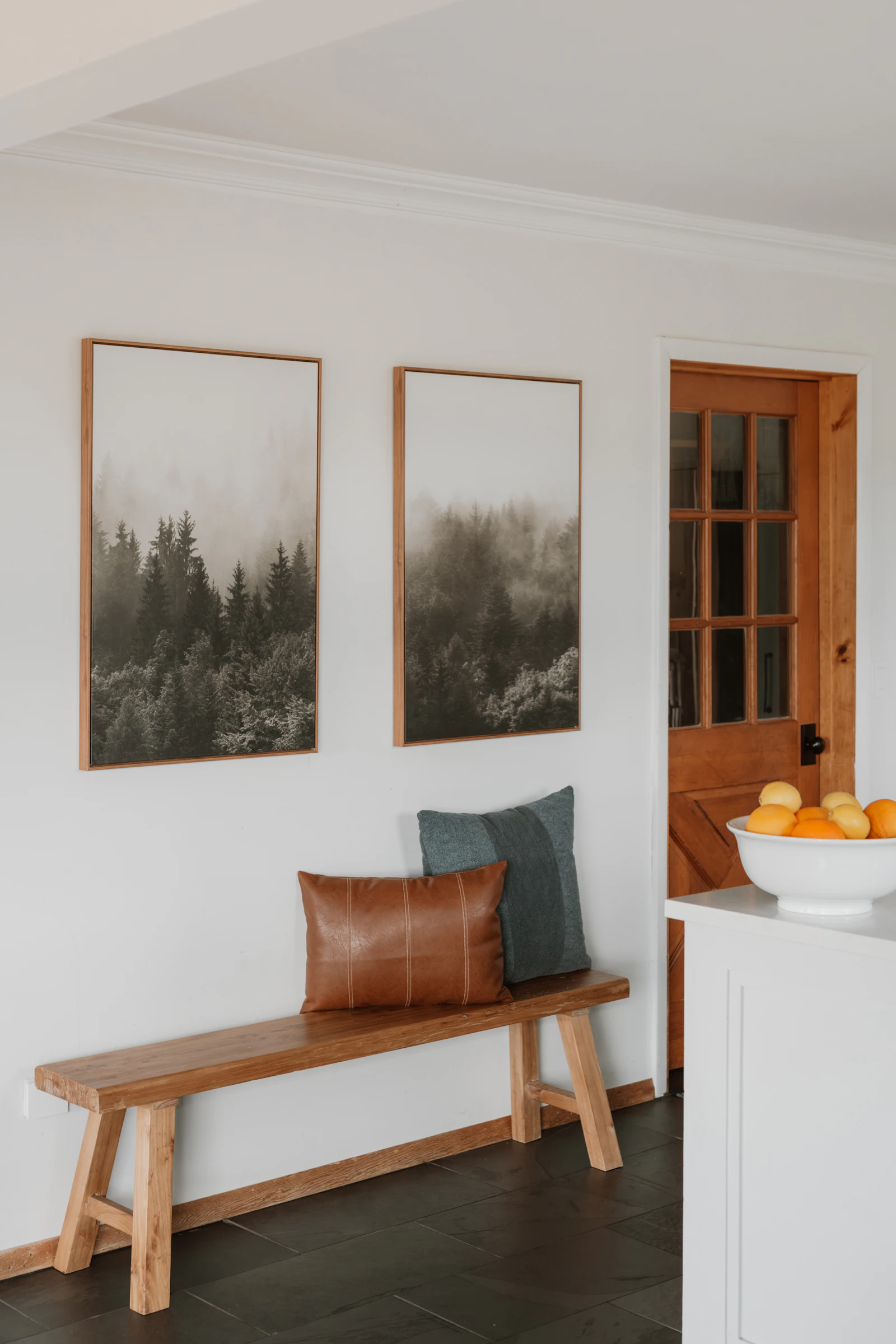 Wooden bench with a leather and a grey cushion below two wood-framed misty forest prints, next to a glazed wooden door