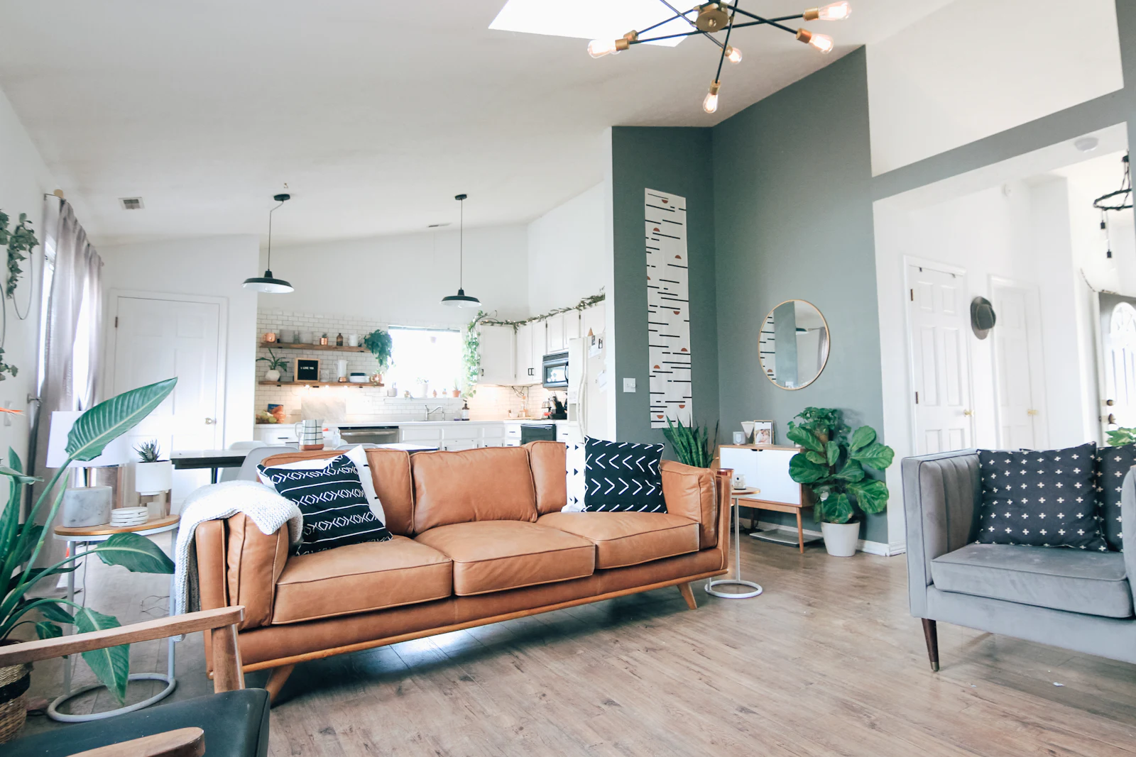 Open-plan living room with a tan leather sofa, a sage-green feature wall, white walls and a kitchen with pendant lights beyond