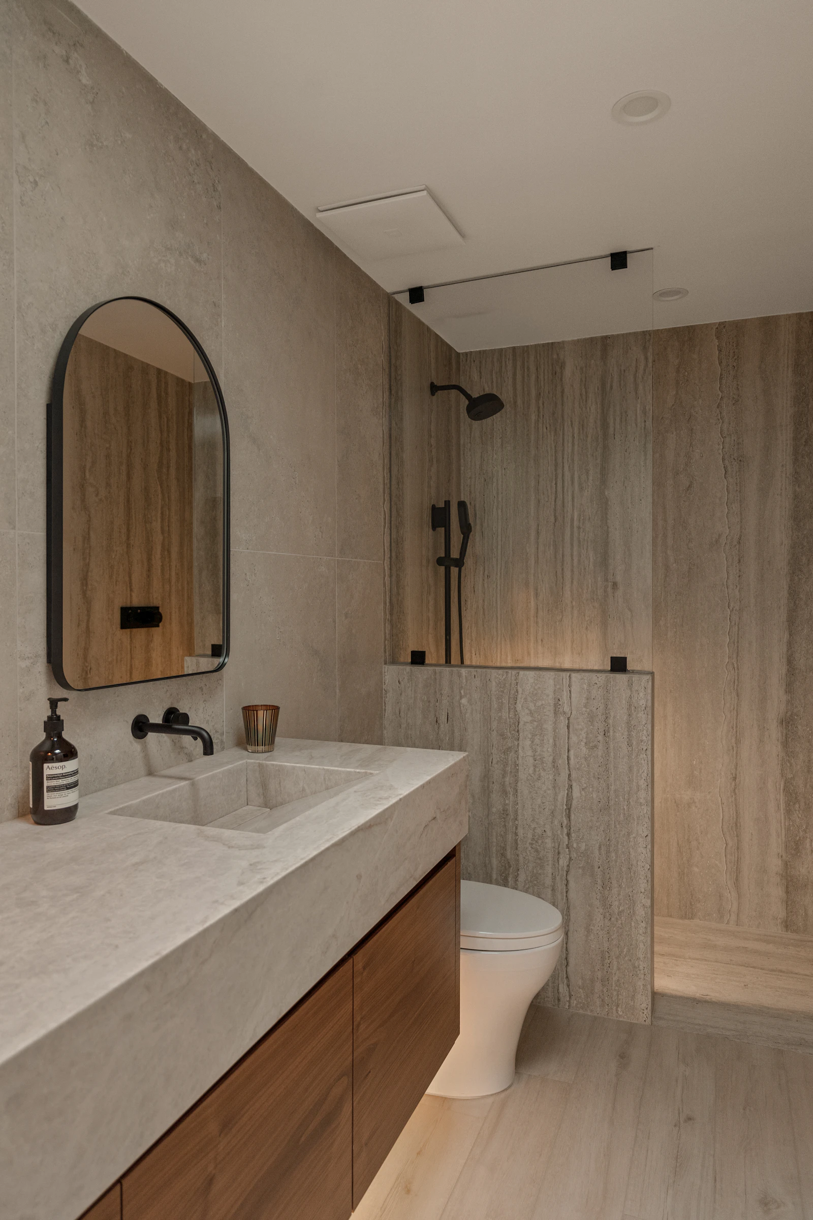 Bathroom with stone-look tiles, a wooden vanity, a round mirror and a pendant light